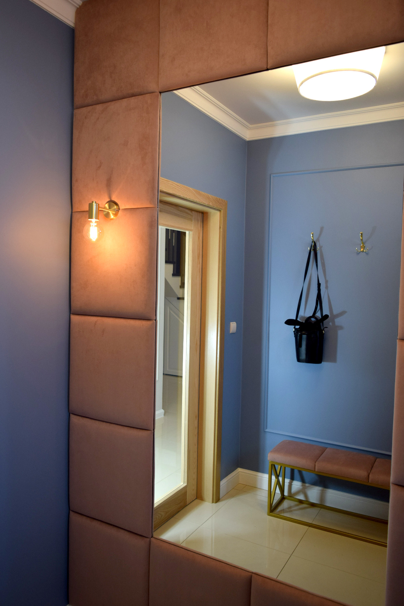 Przedpokój w odcieniach błękitu i pudrowego różu z dużym lustrem w ramie z paneli tapicerowanych, złotym kinkietem i minimalistyczną ławeczką na złotym stelażu, widok na schody przez otwarte drzwi.