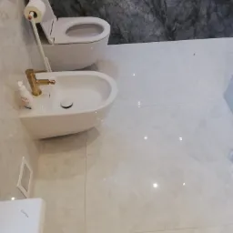 Nowoczesna łazienka z białą ceramiką: toaleta i bidet z armaturą w kolorze złotym, marmurowa ściana w tle, jasne płytki podłogowe.