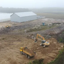 Widok z góry na plac budowy z pracującą koparką i wywrotką załadowaną ziemią, w tle hala magazynowa i szklarnie, teren rozjeżdżony przez maszyny, pochmurny dzień.