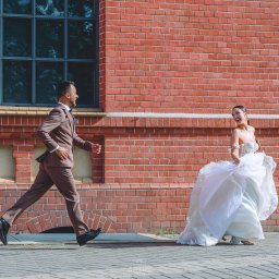 Dron4Pic - Uśmiechnięta para młoda w ruchu: pan młody w brązowym garniturze biegnie, panna młoda w białej sukni ślubnej podnosi spódnicę, na tle ceglanej ściany budynku z oknami.