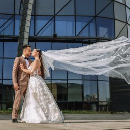 Dron4Pic - Para młoda w objęciach na tle nowoczesnego budynku z ciemnymi oknami; welon powiewa na wietrze, tworząc dramatyczny efekt.