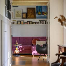 Dron4Pic - Wnętrze mieszkania z fioletową sofą, drewnianą podłogą, dekoracyjną półką nad sofą z obrazami i fotografiami, oraz designerskim stolikiem i krzesłem w stylu retro.