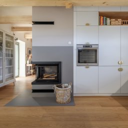 Dron4Pic - Jasne wnętrze salonu z kominkiem i zabudową kuchenną, drewniane belki sufitowe, parkiet w odcieniach miodu, kosz z drewnem obok kominka, nowoczesny design.