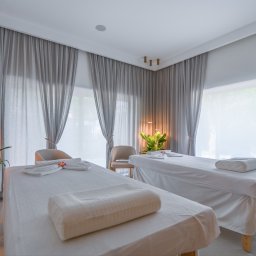 Dron4Pic - Jasne wnętrze gabinetu masażu z dwoma łóżkami, białymi ręcznikami z kwiatami i szarymi zasłonami, oświetlone lampami wiszącymi.