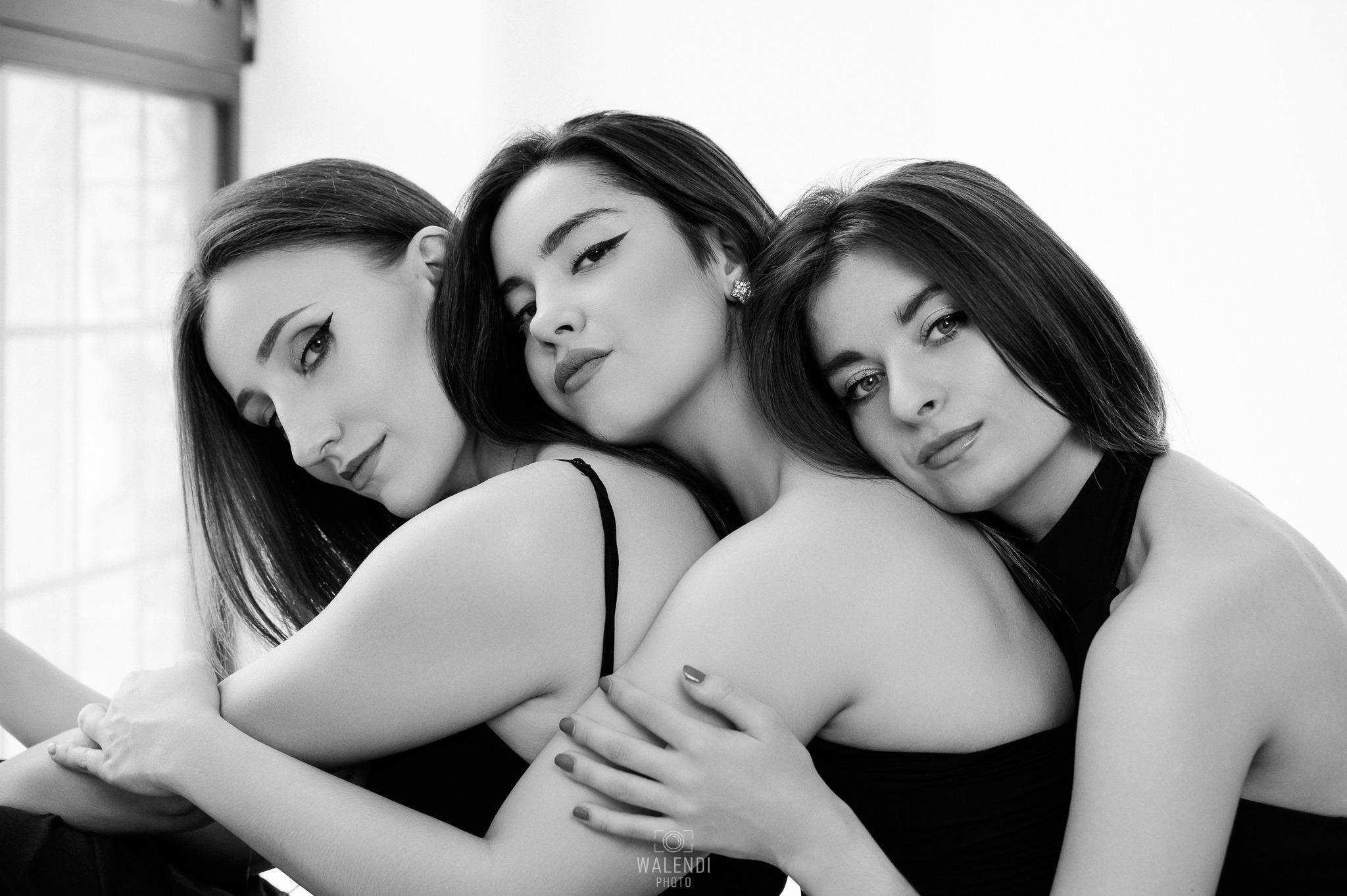 Portret trzech kobiet w czerni, blisko siebie, w czarno-białej tonacji, z wyraźnie zaznaczonym makijażem oczu i subtelną biżuterią, z delikatnym oświetleniem.