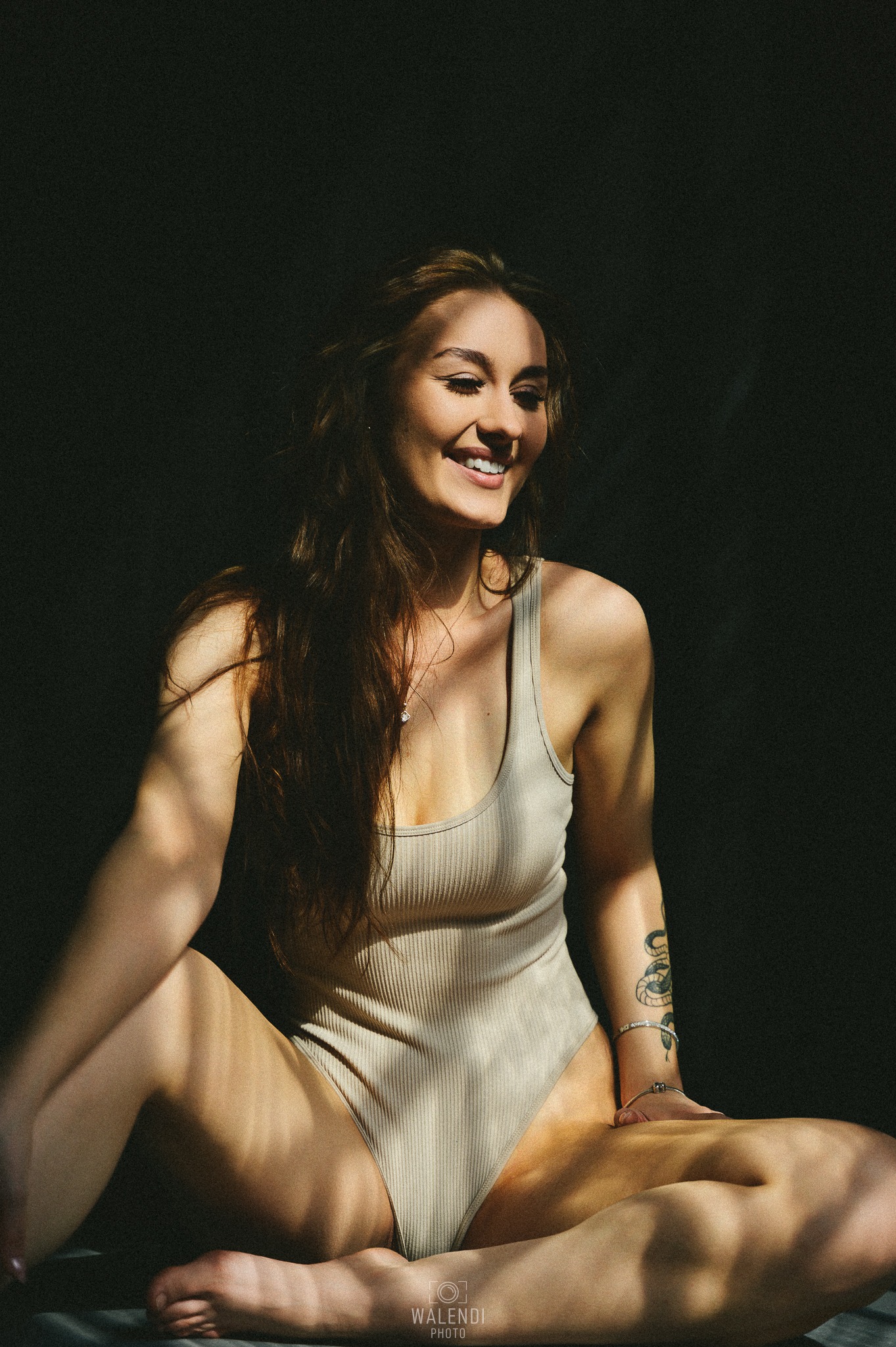 Uśmiechnięta brunetka w beżowym body siedzi w kontraście światła i cienia, z widocznym tatuażem węża na ramieniu i delikatnym naszyjnikiem z wisiorkiem.