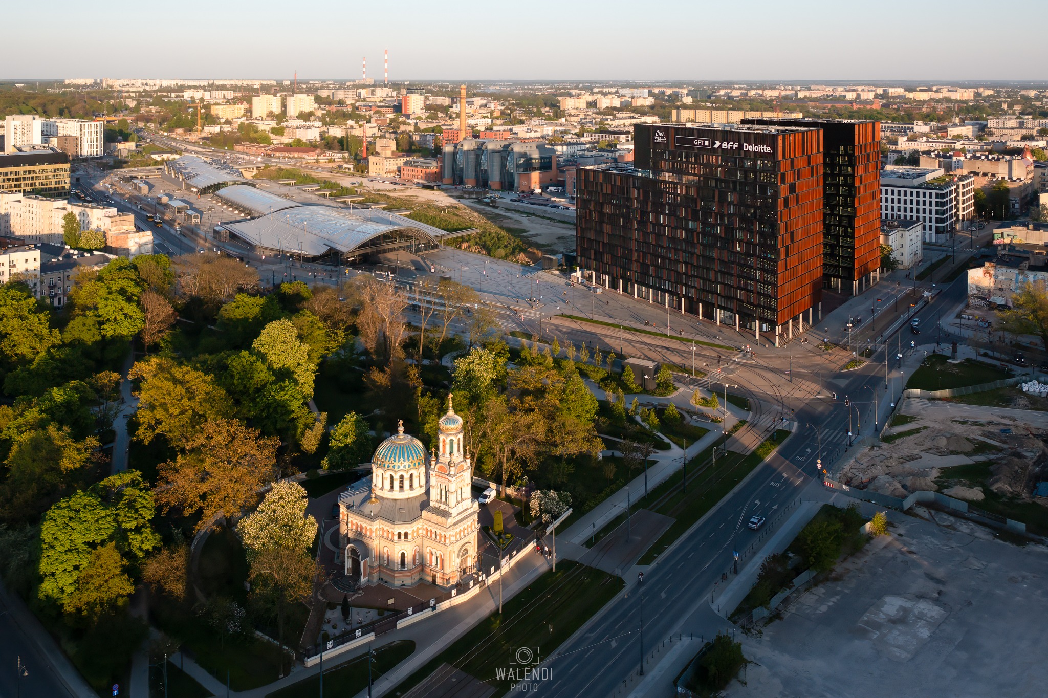 Panorama miasta z lotu ptaka ukazuje cerkiew z błękitnymi kopułami, nowoczesny dworzec kolejowy, biurowce z logo firm i kominy w tle, wszystko oświetlone ciepłym, popołudniowym światłem.