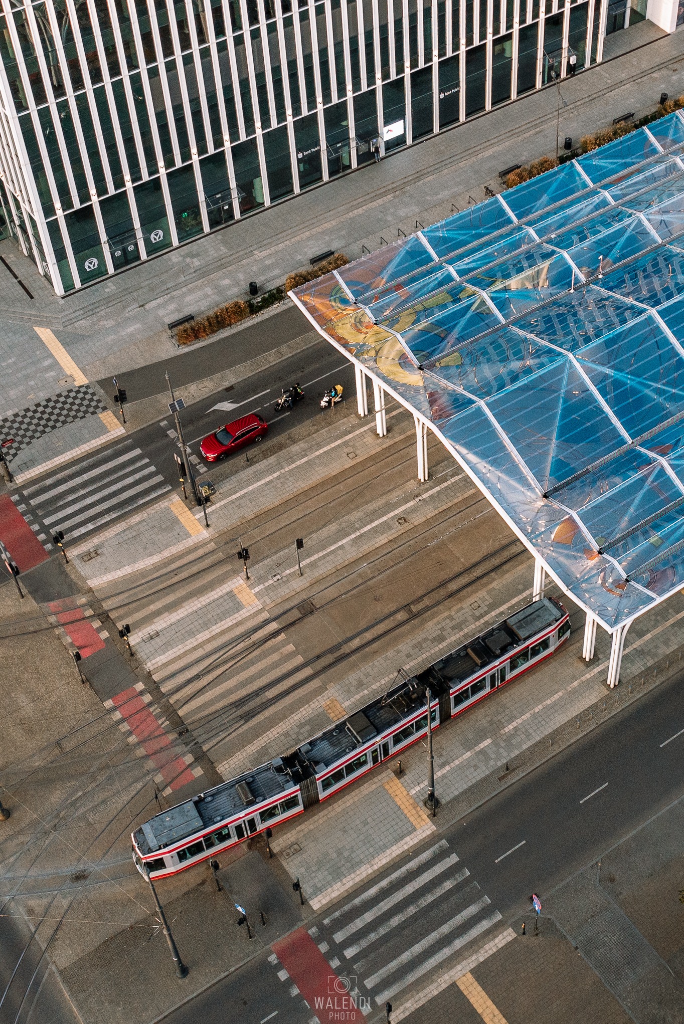 Widok z góry na przystanek tramwajowy z przezroczystym zadaszeniem z motywami graficznymi, czerwony samochód na przejściu dla pieszych, biały budynek biurowy z logo banku w tle.