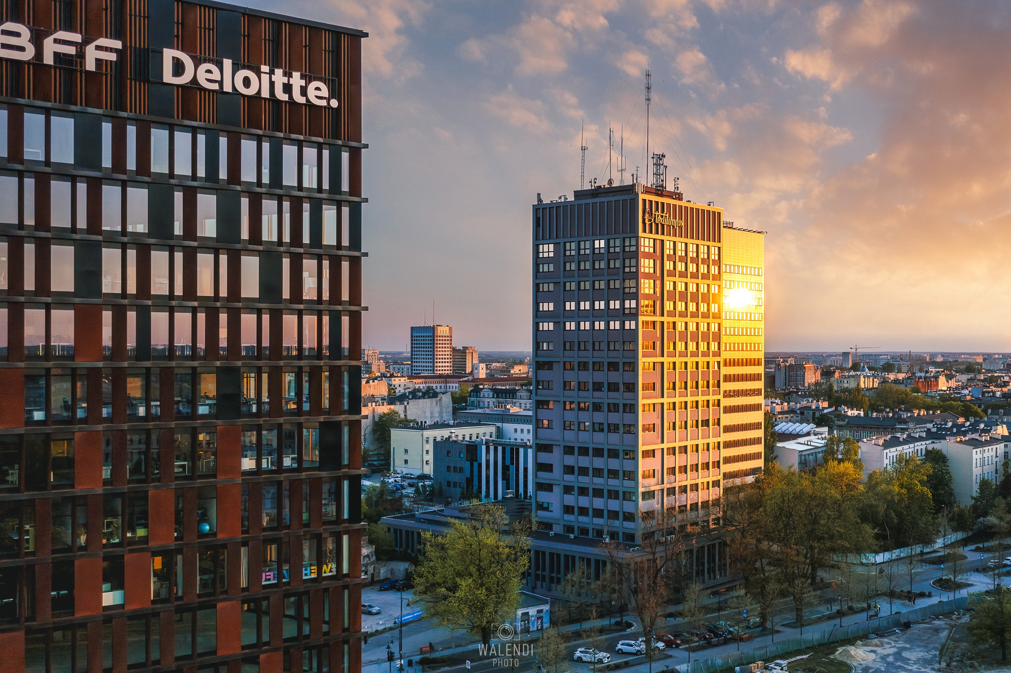 Panorama miasta z nowoczesnymi biurowcami o zachodzie słońca, widoczne logo firm Bff Deloitte i Textilimpex na budynkach.