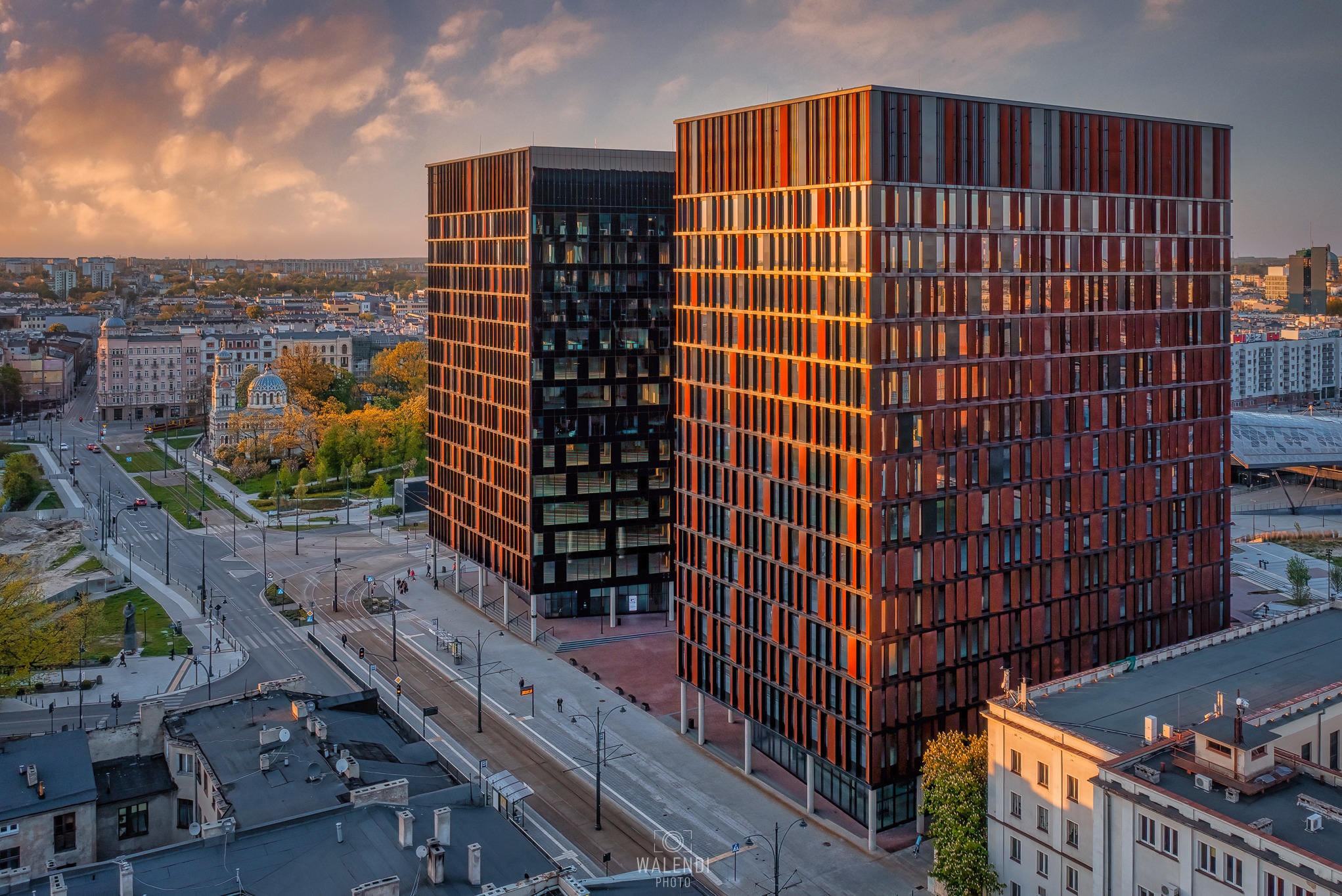 Panorama miasta z dwoma nowoczesnymi biurowcami o pomarańczowo-brązowej fasadzie w promieniach zachodzącego słońca, widok z góry na ulicę i zabytkową architekturę w tle.