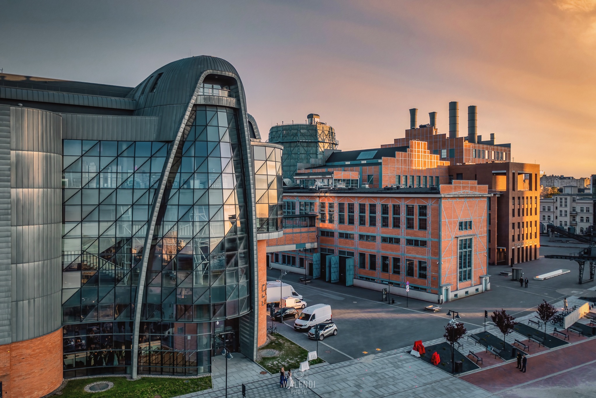 Kompleks budynków pofabrycznych z nowoczesną przeszkloną konstrukcją, oświetlonych ciepłym światłem zachodzącego słońca, widok z góry na plac z zaparkowanymi samochodami i spacerującymi ludźmi.