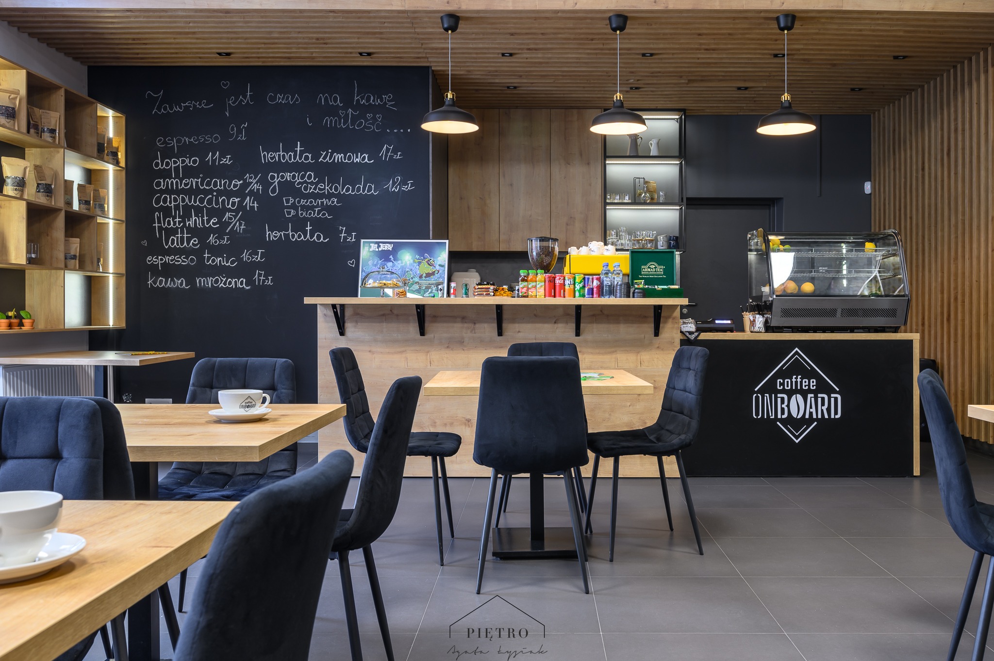Wnętrze kawiarni z drewnianymi stołami i krzesłami obitymi granatowym aksamitem, czarna ściana z menu napisanym kredą, oświetlenie punktowe i logo 'Coffee Onboard' na barze.