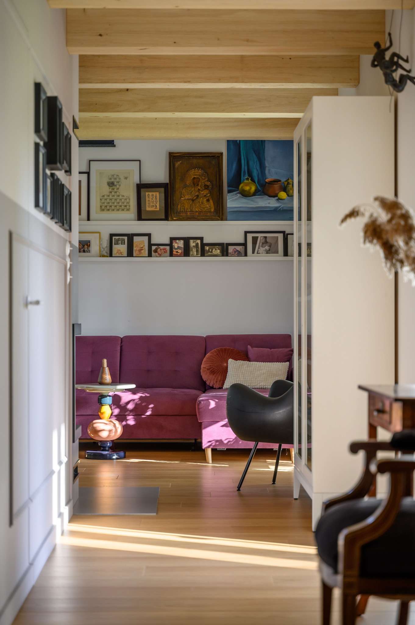 Wnętrze mieszkania z fioletową sofą, drewnianą podłogą, dekoracyjną półką nad sofą z obrazami i fotografiami, oraz designerskim stolikiem i krzesłem w stylu retro.