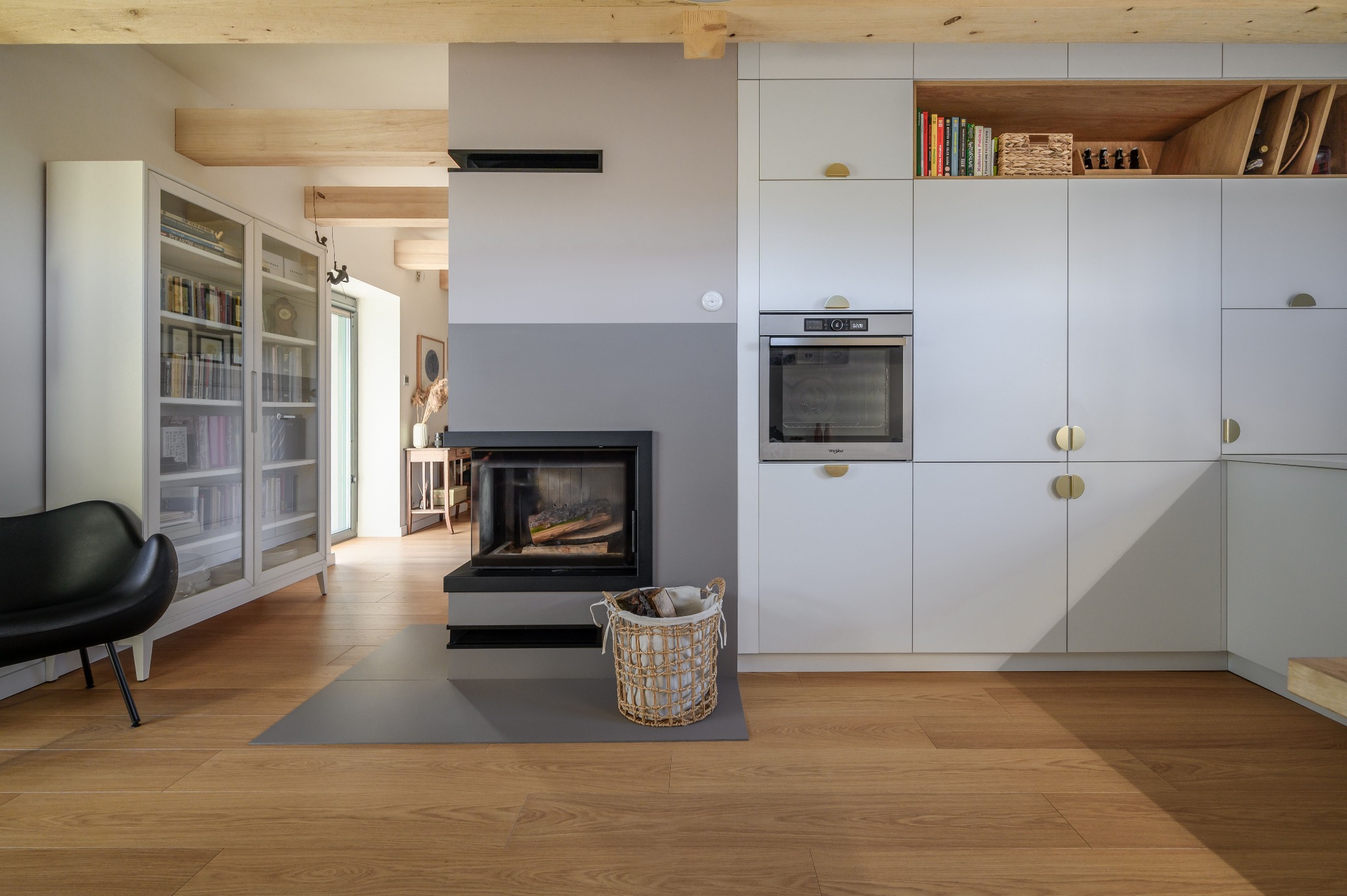 Jasne wnętrze salonu z kominkiem i zabudową kuchenną, drewniane belki sufitowe, parkiet w odcieniach miodu, kosz z drewnem obok kominka, nowoczesny design.