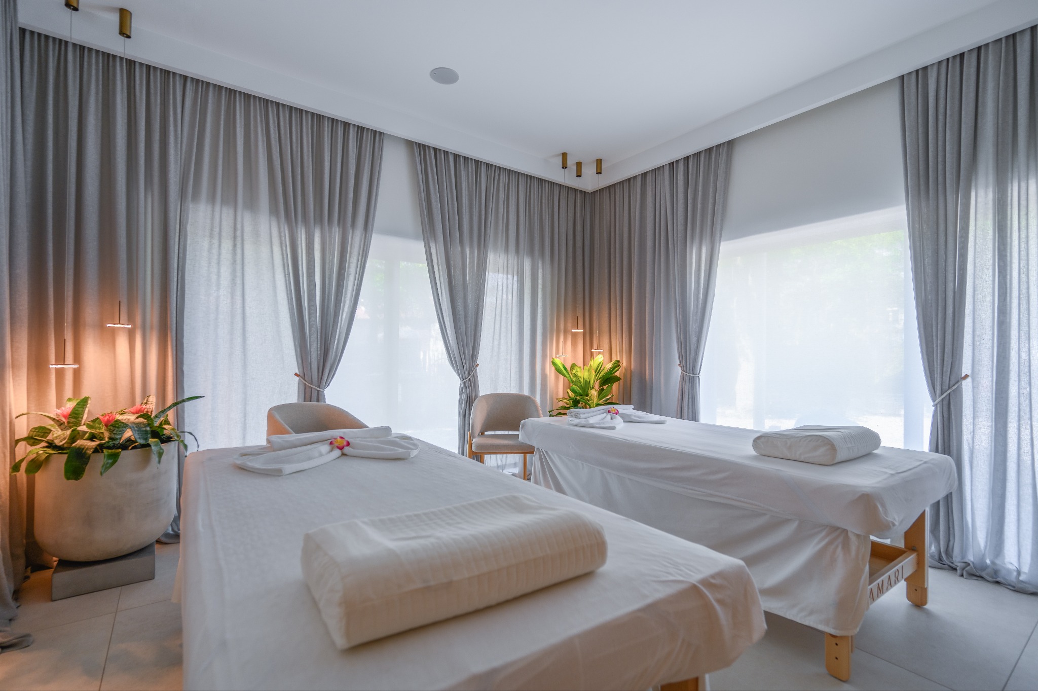 Jasne wnętrze gabinetu masażu z dwoma łóżkami, białymi ręcznikami z kwiatami i szarymi zasłonami, oświetlone lampami wiszącymi.