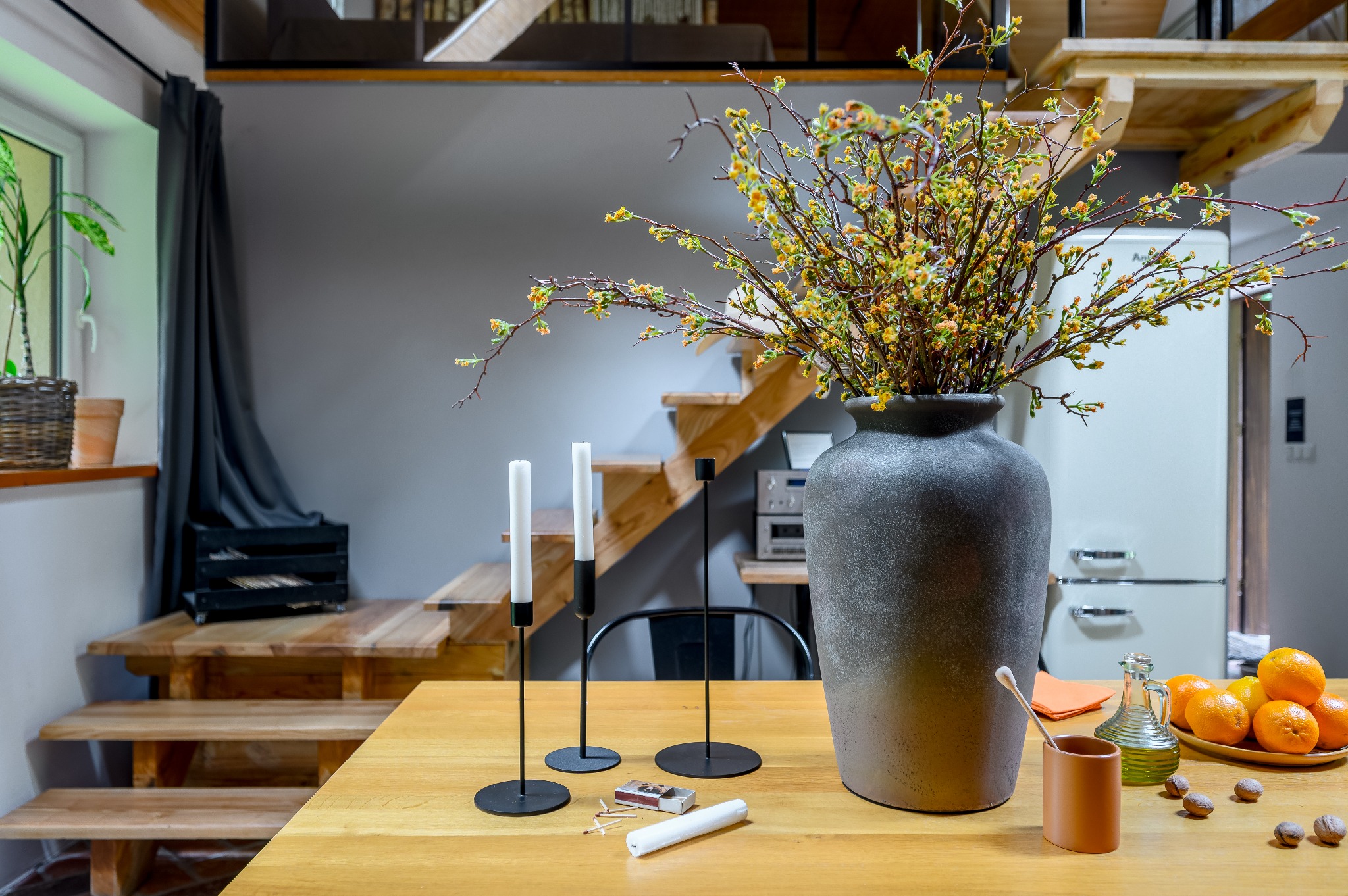 Stylowa aranżacja wnętrza z drewnianym stołem, na którym znajdują się świece w czarnych świecznikach, zapałki, duży szary wazon z kwitnącymi gałązkami, naczynie z pomarańczami i butelka oliwy;...