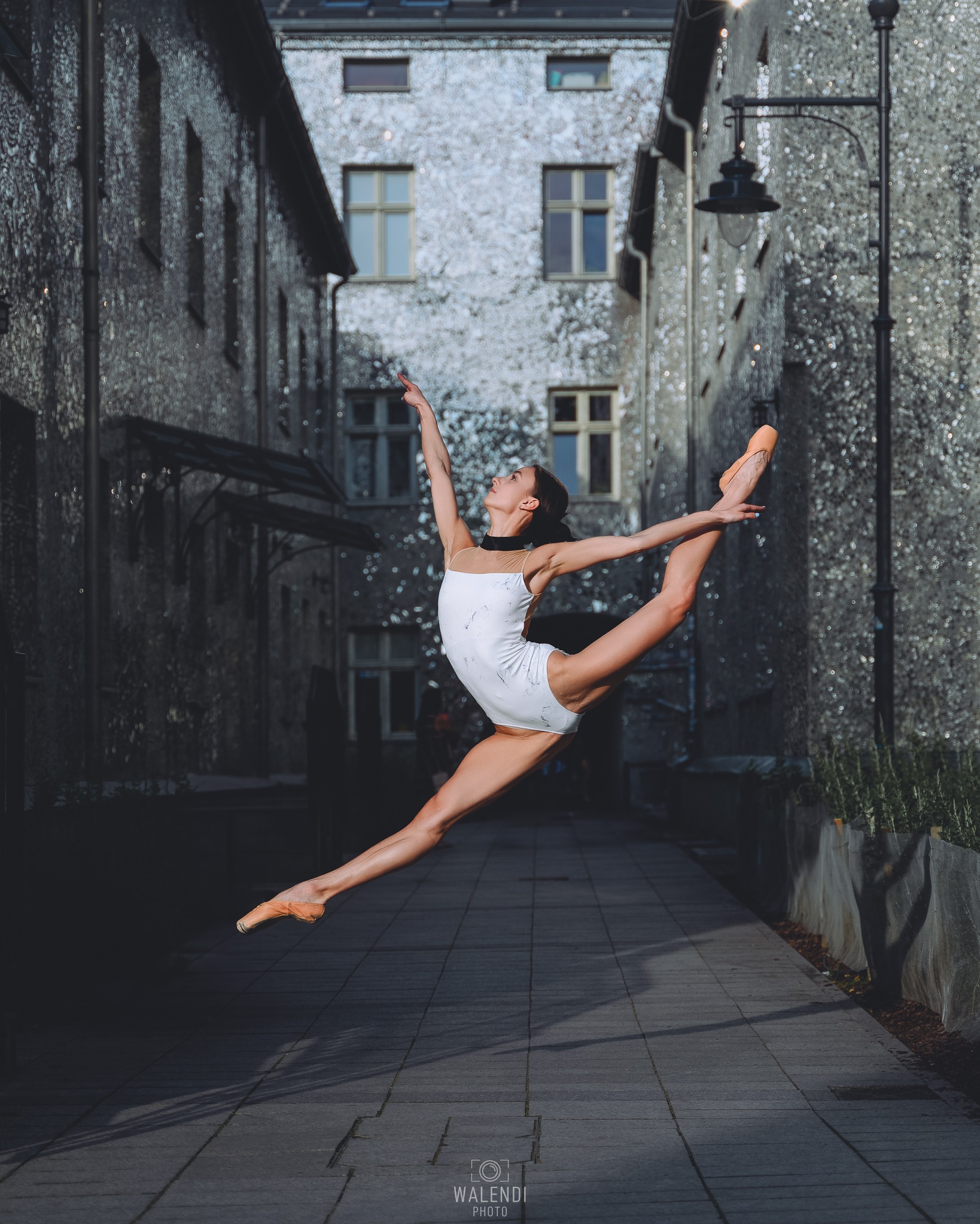 Baletnica w białym stroju wykonuje szpagat w powietrzu na tle budynków z lustrzaną elewacją i brukowanej uliczki.