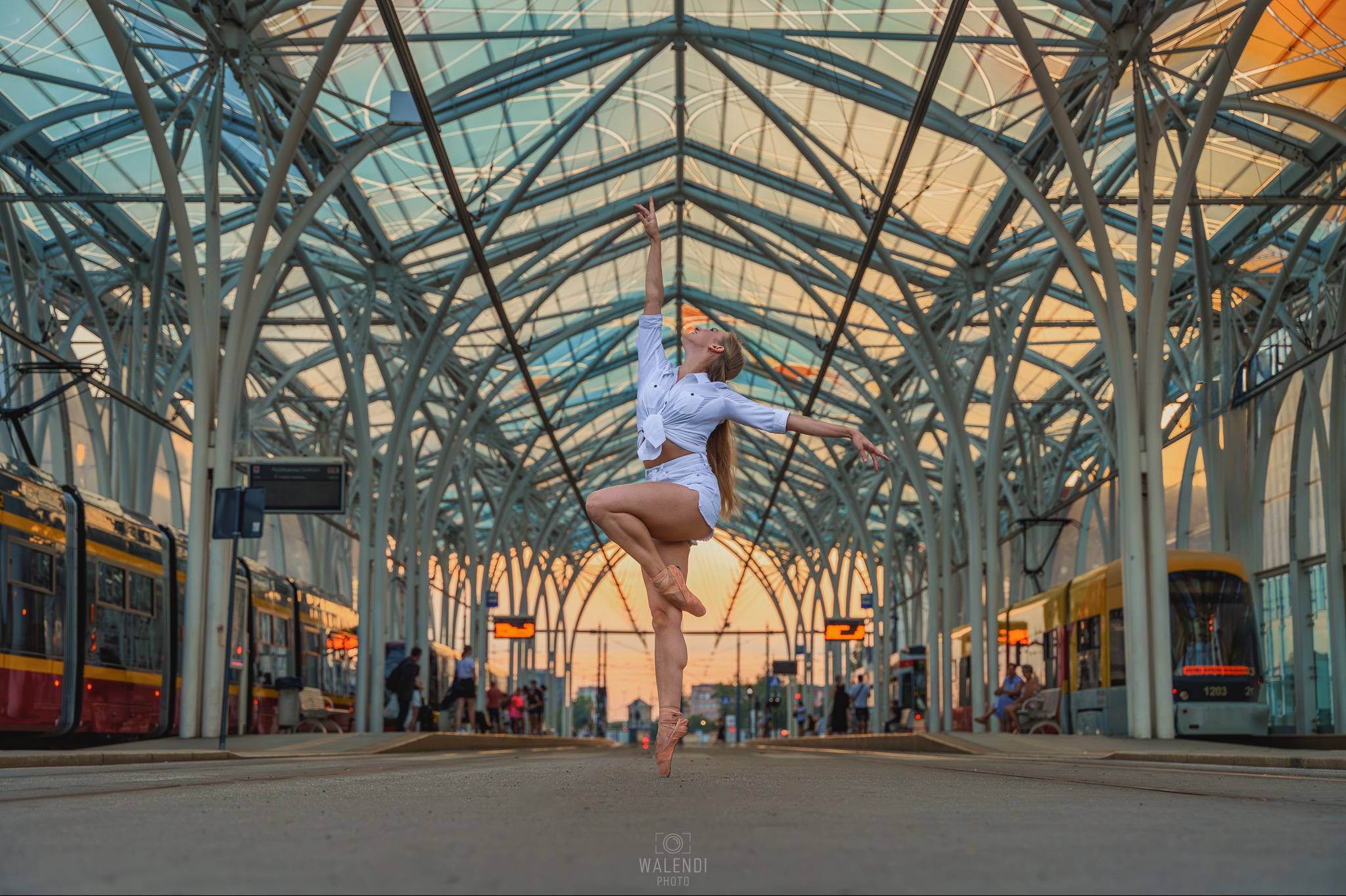 Baletnica w białym stroju pozuje na tle futurystycznej konstrukcji przystanku tramwajowego w Aleksandrowie Łódzkim, z tramwajami po bokach.