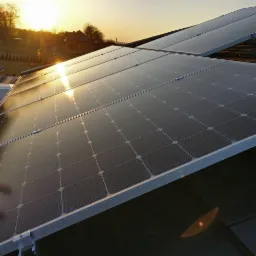 Panele słoneczne na dachu domu oświetlone promieniami zachodzącego słońca, widok na okoliczne budynki i drzewa w tle.