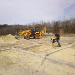 Prace ziemne pod fundamenty domu: zagęszczanie piasku ubijarką wibracyjną, w tle koparka JCB i stos palet.
