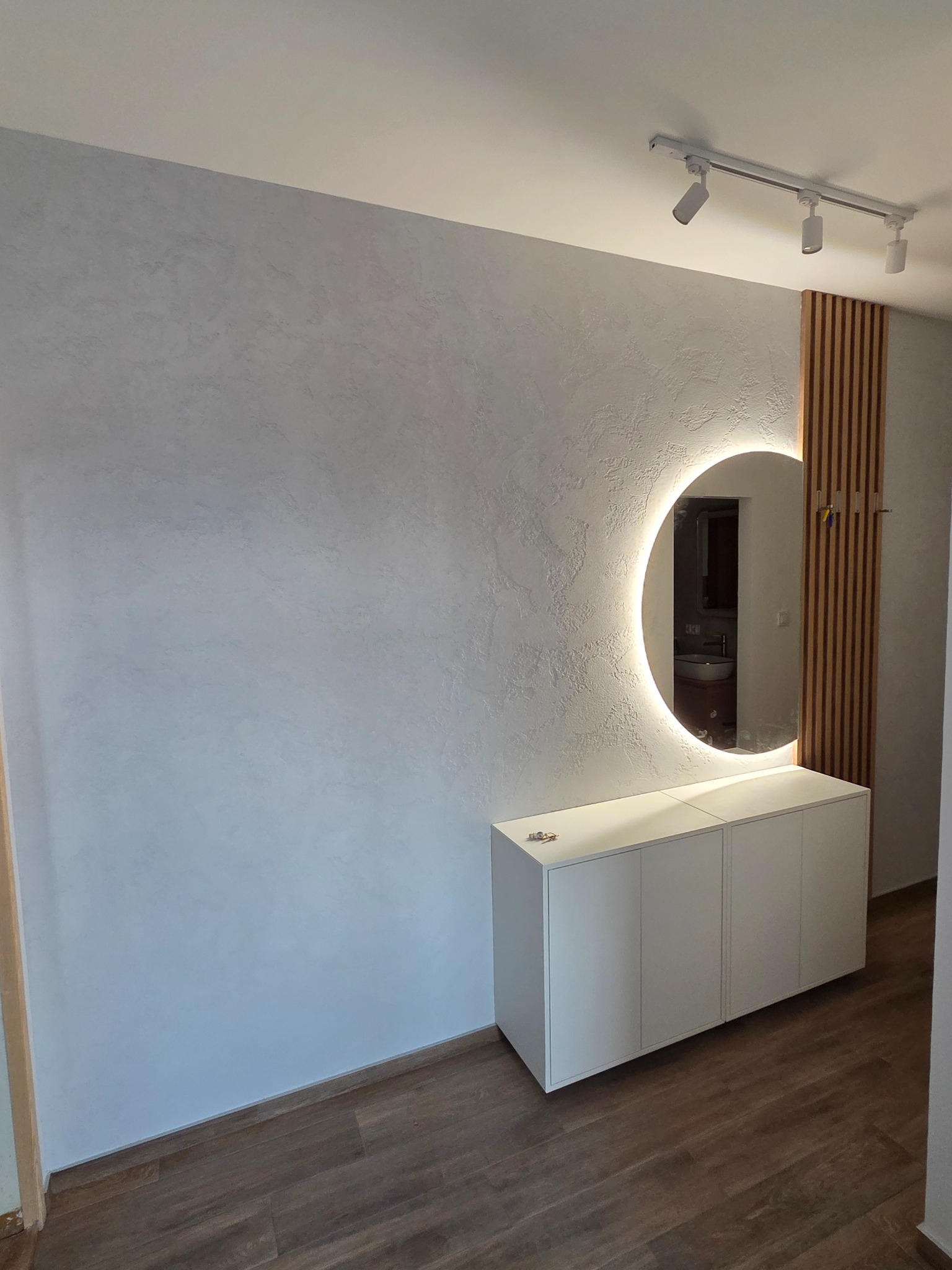 Wykończenie przedpokoju: dekoracyjna ściana z efektem stiuku, podświetlane okrągłe lustro nad białą szafką, drewniane lamele i parkiet. Nowoczesny design.