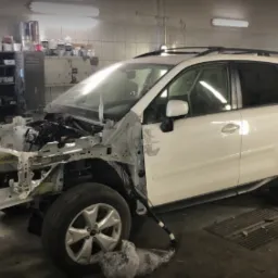 Biały samochód osobowy marki Subaru Forester w trakcie naprawy blacharskiej, z częściowo zdemontowanym przodem, widoczne elementy konstrukcyjne i zawieszenie, w tle warsztat z regałami i narzędziami.