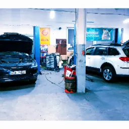 Wnętrze warsztatu samochodowego z dwoma autami: czarnym sedanem z otwartą maską i białym kombi z otwartą klapą bagażnika, widoczny wózek narzędziowy i beczka na olej.