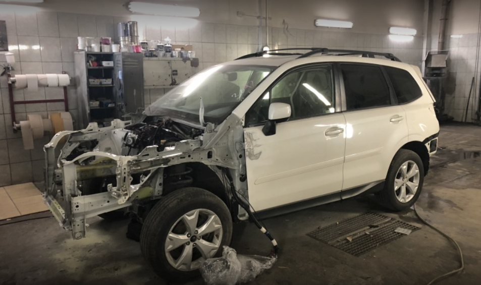 Biały samochód osobowy marki Subaru Forester w trakcie naprawy blacharskiej, z częściowo zdemontowanym przodem, widoczne elementy konstrukcyjne i zawieszenie, w tle warsztat z regałami i narzędziami.