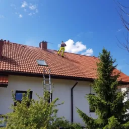 Pracownik w żółtych spodniach i szarej kamizelce, z myjką ciśnieniową na dachu domu z czerwoną dachówką, obok komin i drabina, błękitne niebo z chmurami w tle.