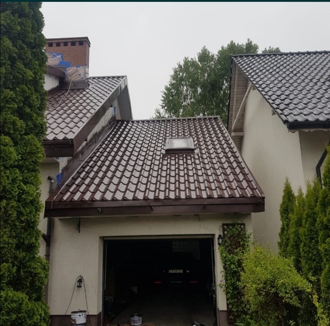 Dom z brązowym dachem z dachówki, widoczne rynny, okno dachowe i garaż z otwartą bramą.