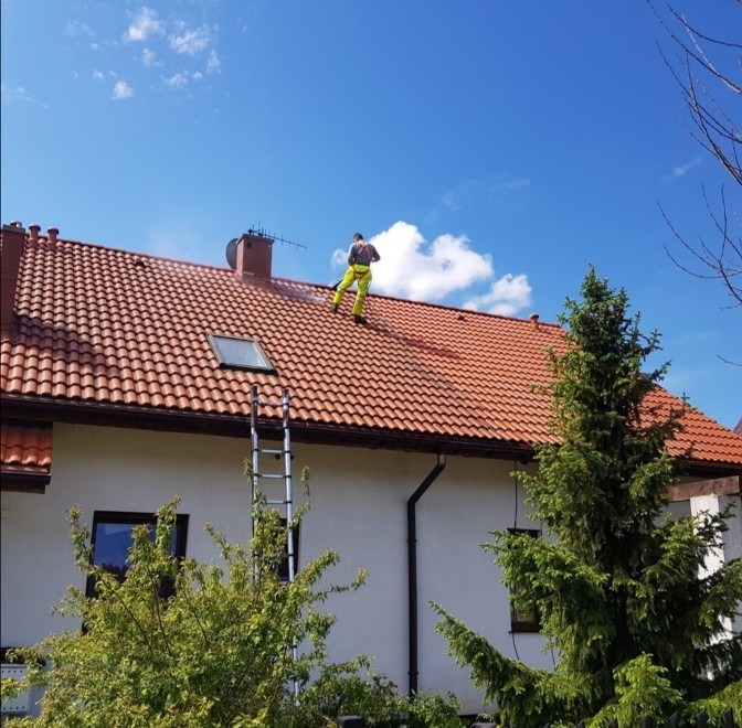 Pracownik w żółtych spodniach i szarej kamizelce, z myjką ciśnieniową na dachu domu z czerwoną dachówką, obok komin i drabina, błękitne niebo z chmurami w tle.