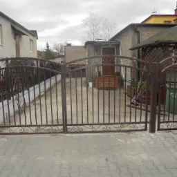 Kuta, brązowa brama wjazdowa z furtką, ozdobiona wzorem z motywem ślimaków, prowadząca na posesję z kostki brukowej, widok z zewnątrz.