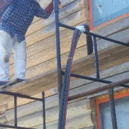 Pracownik w kombinezonie ochronnym i masce na rusztowaniu, czyści drewnianą elewację domu z bali za pomocą specjalistycznego sprzętu. Okna zabezpieczone folią.