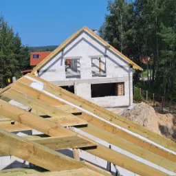 Widok na budowę domu murowanego z perspektywy drewnianej konstrukcji dachu, w tle widać las i błękitne niebo.