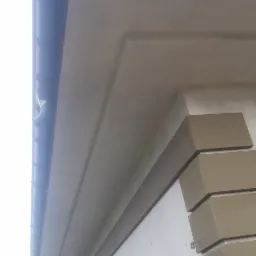 Fragment elewacji domu z pomalowanymi elementami dekoracyjnymi w odcieniach beżu i szarości, widoczny fragment rynny i dach sąsiedniego budynku.