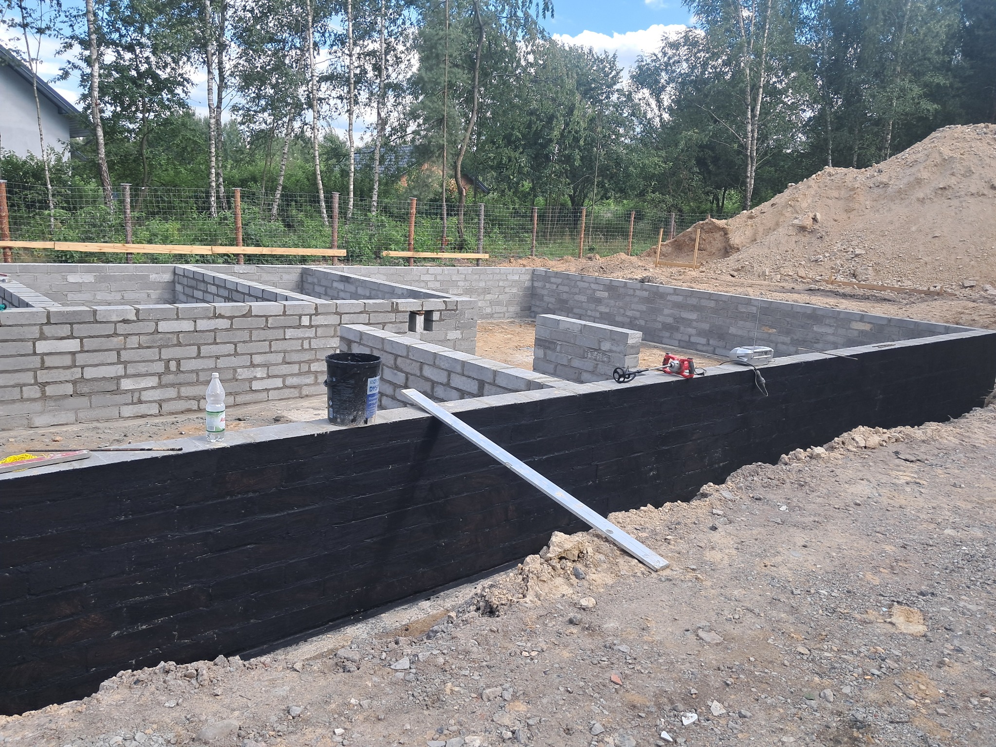 Budowa fundamentów domu z bloczków betonowych, zewnętrzna ściana fundamentowa zabezpieczona czarną izolacją, poziomica oparta o ścianę, w tle hałda piachu i drzewa.