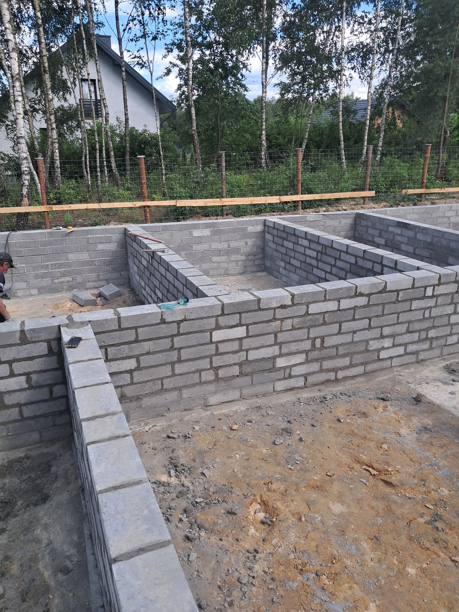 Wznoszenie fundamentów z szarego bloczka betonowego, widoczne zbrojenie, w tle drzewa i fragment domu jednorodzinnego.