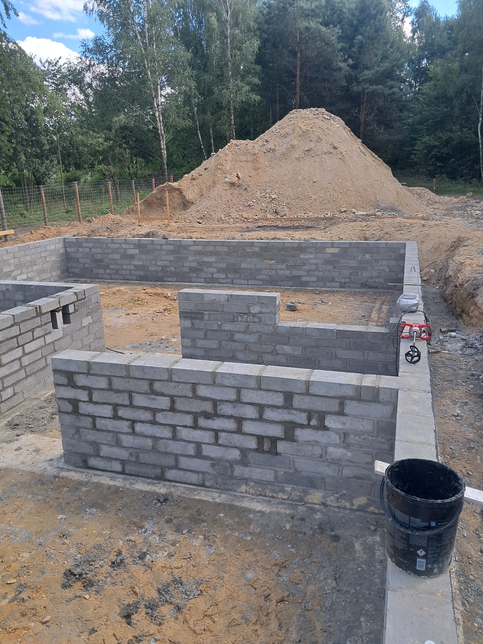 Wznoszenie fundamentów z bloczków betonowych na placu budowy, widoczny mikser do zaprawy, wiadro i pryzma piasku w tle.