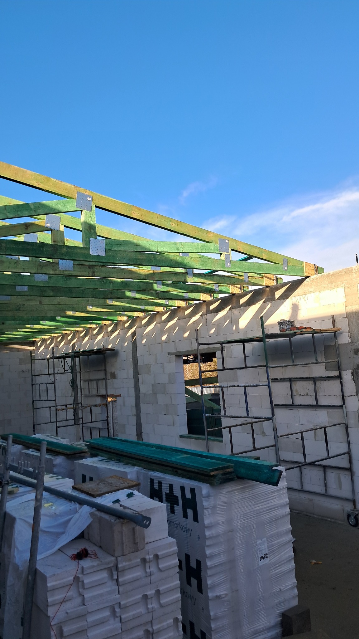 Wznoszenie ścian domu z bloczków H+H, widoczne rusztowania i drewniana konstrukcja dachu z metalowymi łącznikami na tle błękitnego nieba.