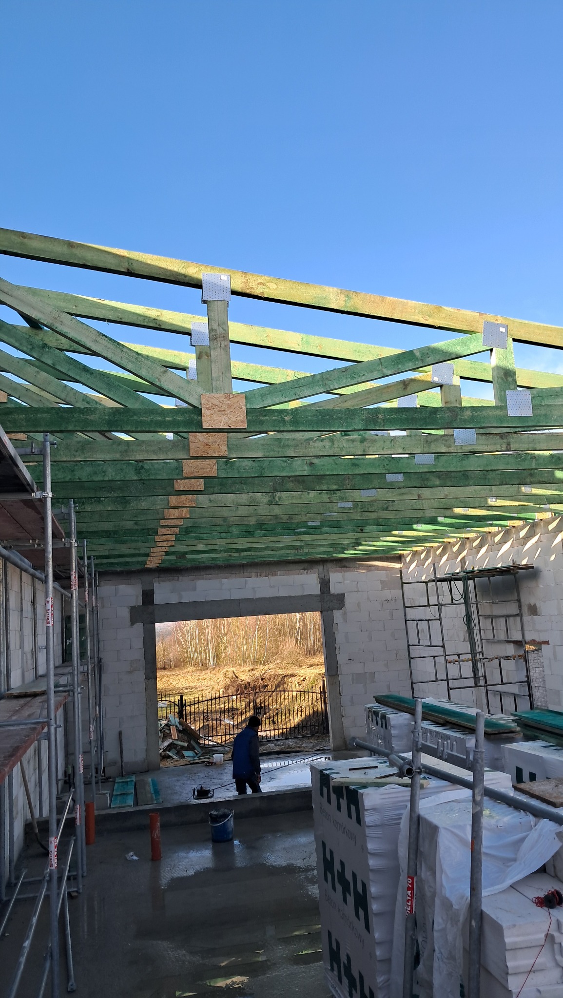 Wnętrze domu w trakcie budowy: widoczne ściany z bloczków, drewniana konstrukcja dachu z impregnacją na zielono, rusztowania i materiały budowlane H+H, zarys sylwetki pracownika w oddali.