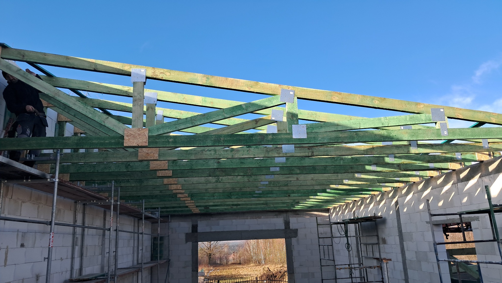 Konstrukcja dachu z zielonych belek drewnianych połączonych metalowymi płytkami, widok od wewnątrz surowego budynku z białych pustaków, na pierwszym planie rusztowania.