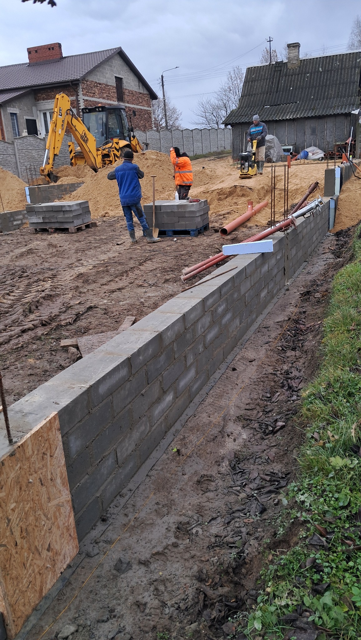 Budowa fundamentów domu: wznoszenie muru z bloczków betonowych, koparka w tle, widoczni pracownicy i przygotowany teren pod budowę.