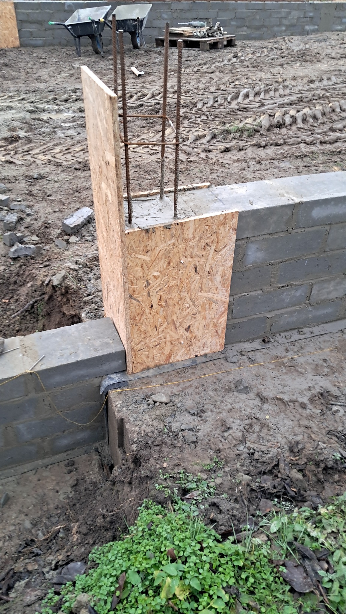 Wznoszenie fundamentów domu z bloczków betonowych, widoczny szalunek z płyty OSB dla słupa zbrojeniowego oraz dwa wózki budowlane w tle.