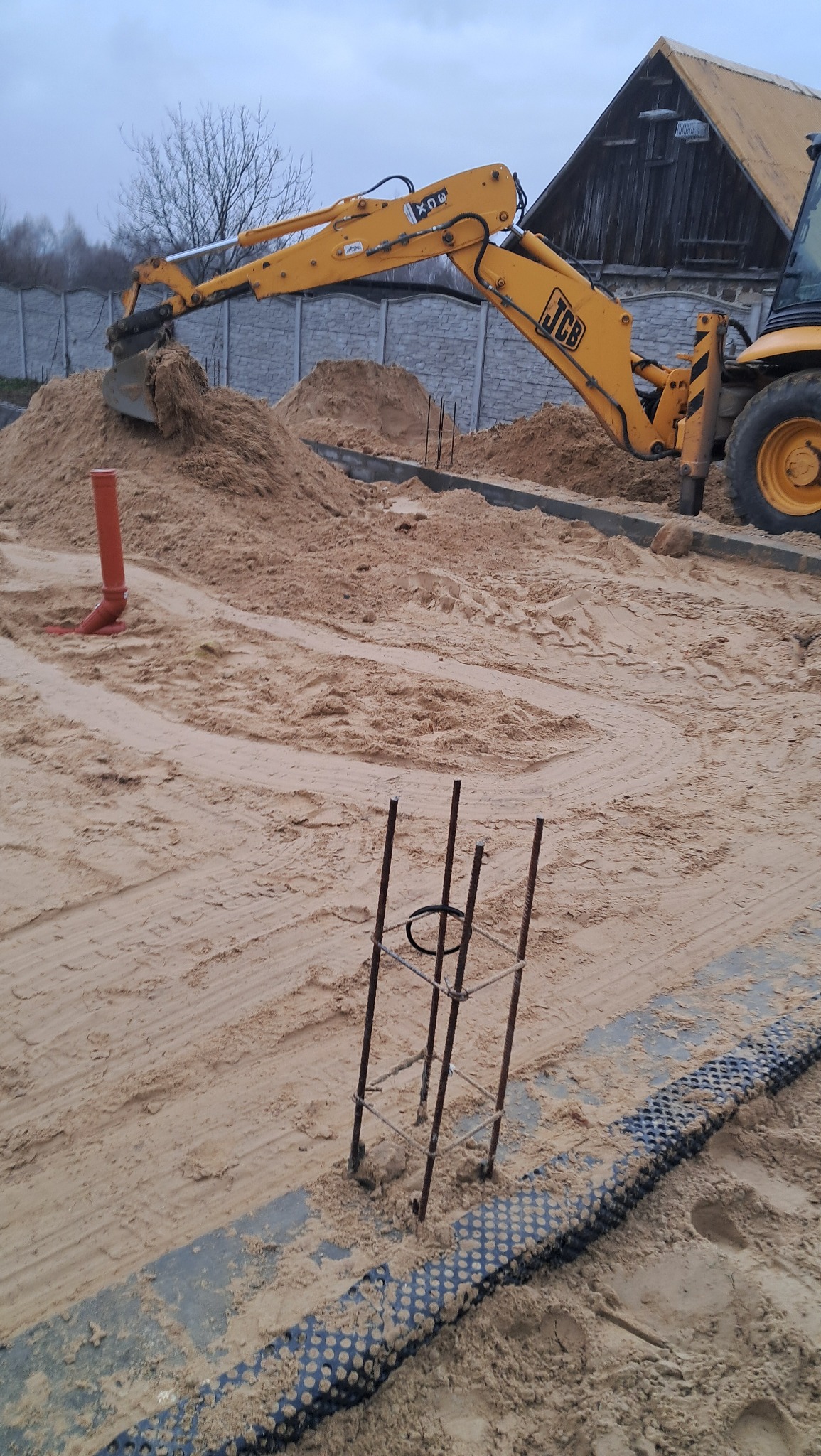 Żółta koparko-ładowarka JCB zasypuje fundamenty piaskiem, widoczne zbrojenia i rura kanalizacyjna, w tle betonowe ogrodzenie i budynek gospodarczy.