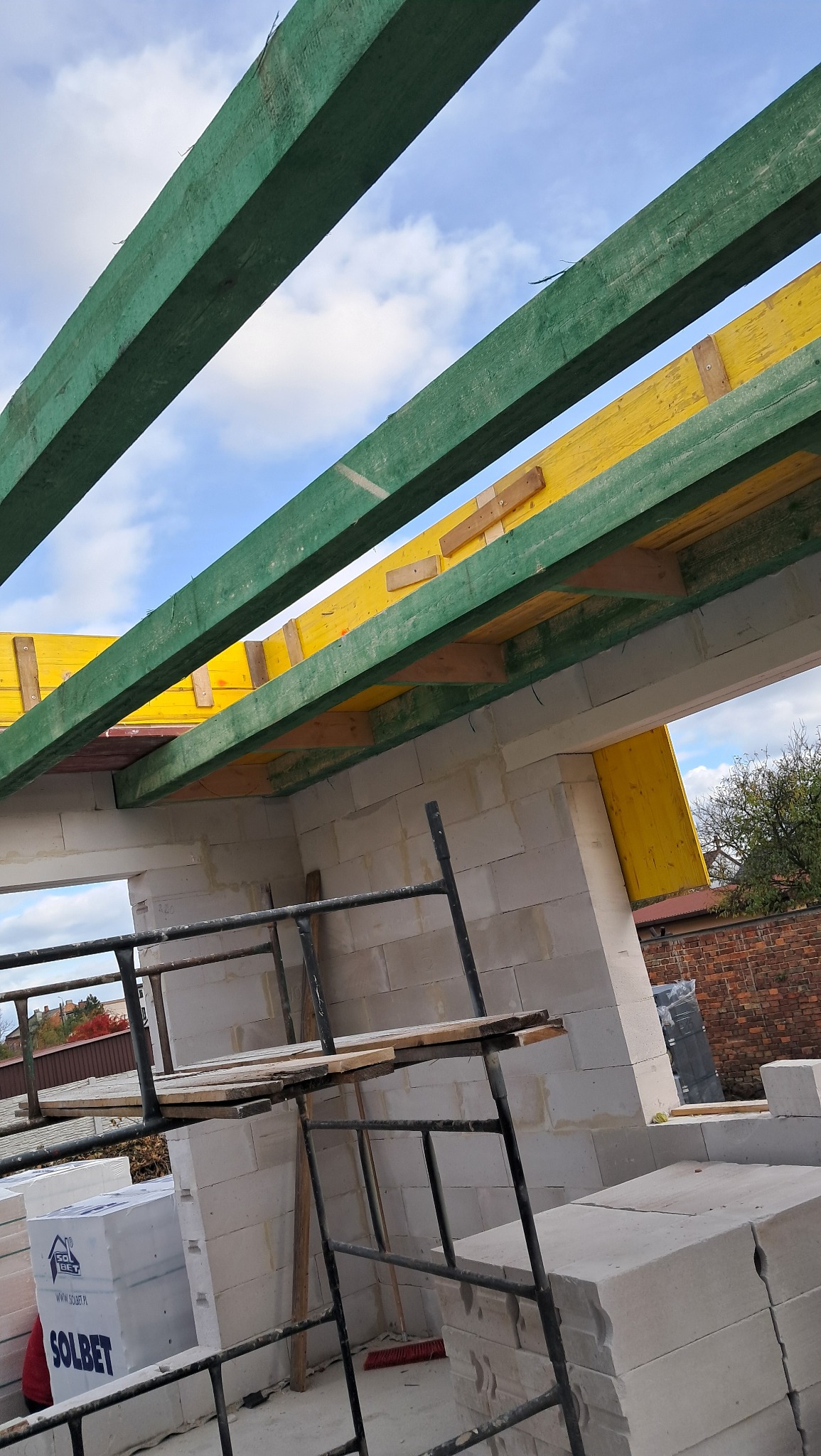 Wznoszenie domu murowanego z bloczków betonu komórkowego, widoczne zielone i żółte elementy konstrukcji dachu oraz rusztowanie.