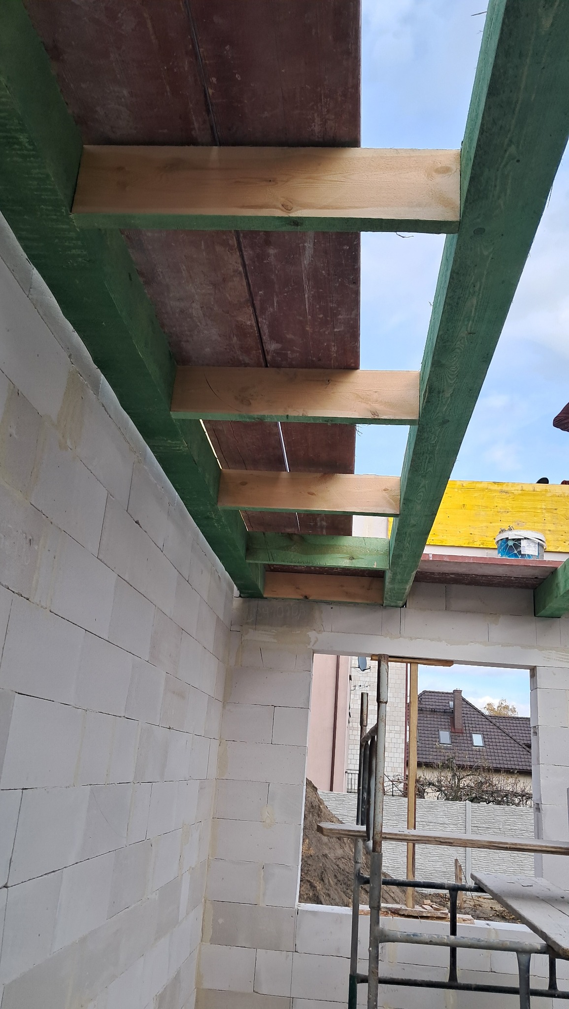 Wnętrze domu w trakcie budowy, widoczne ściany z białych bloczków, drewniana konstrukcja dachu z zielonymi belkami i częściowo ułożonymi deskami, przez okno widać budynki w oddali i rusztowanie...