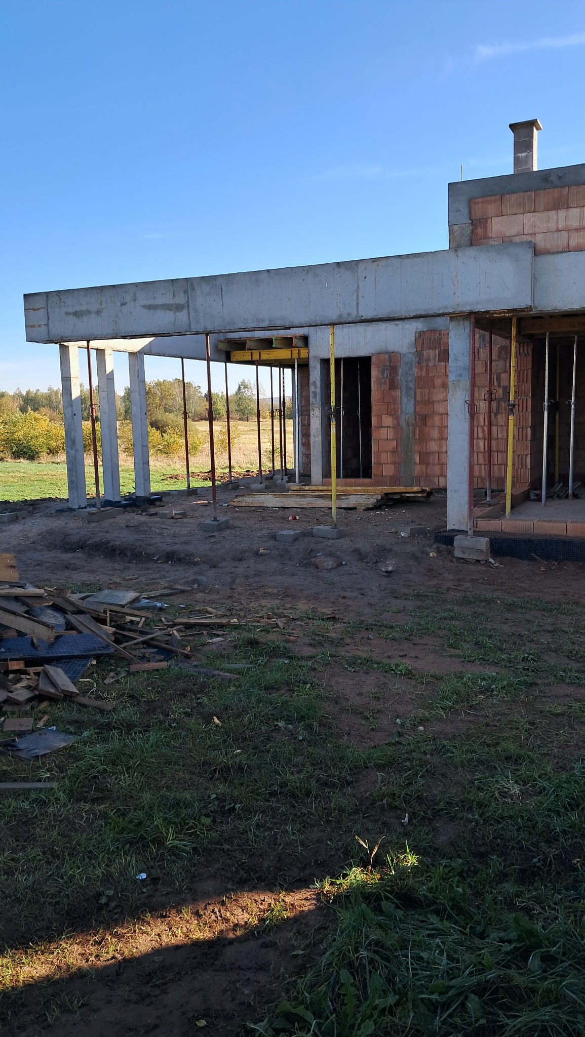 Surowy stan budowy domu z czerwonej cegły, widoczne betonowe filary, żółte podpory i prowizoryczne deskowanie, w tle jesienny krajobraz.