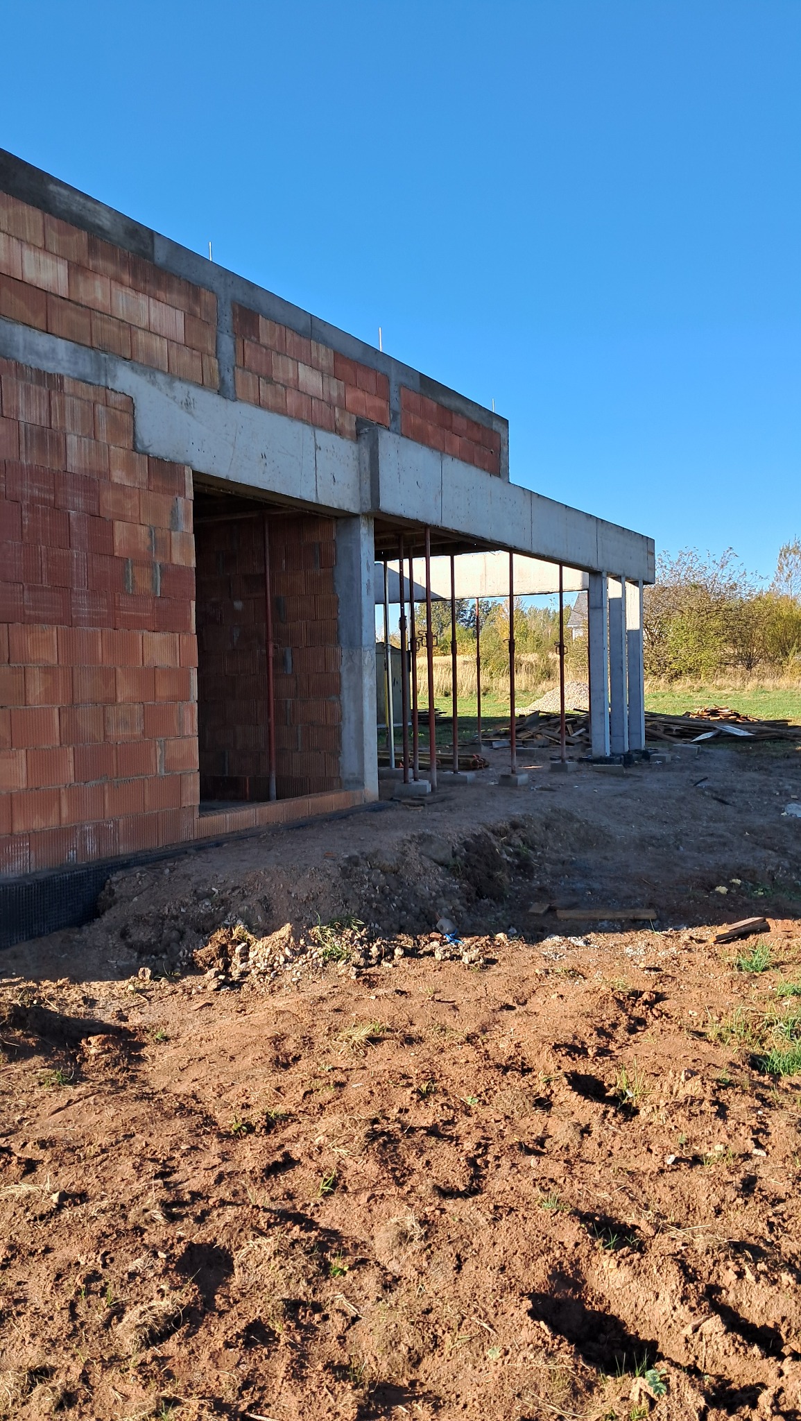 Surowy stan budowy domu z czerwonej cegły, widoczne betonowe nadproża i filary podtrzymujące strop, teren wokół w stanie ziemnym, w tle zieleń i błękitne niebo.