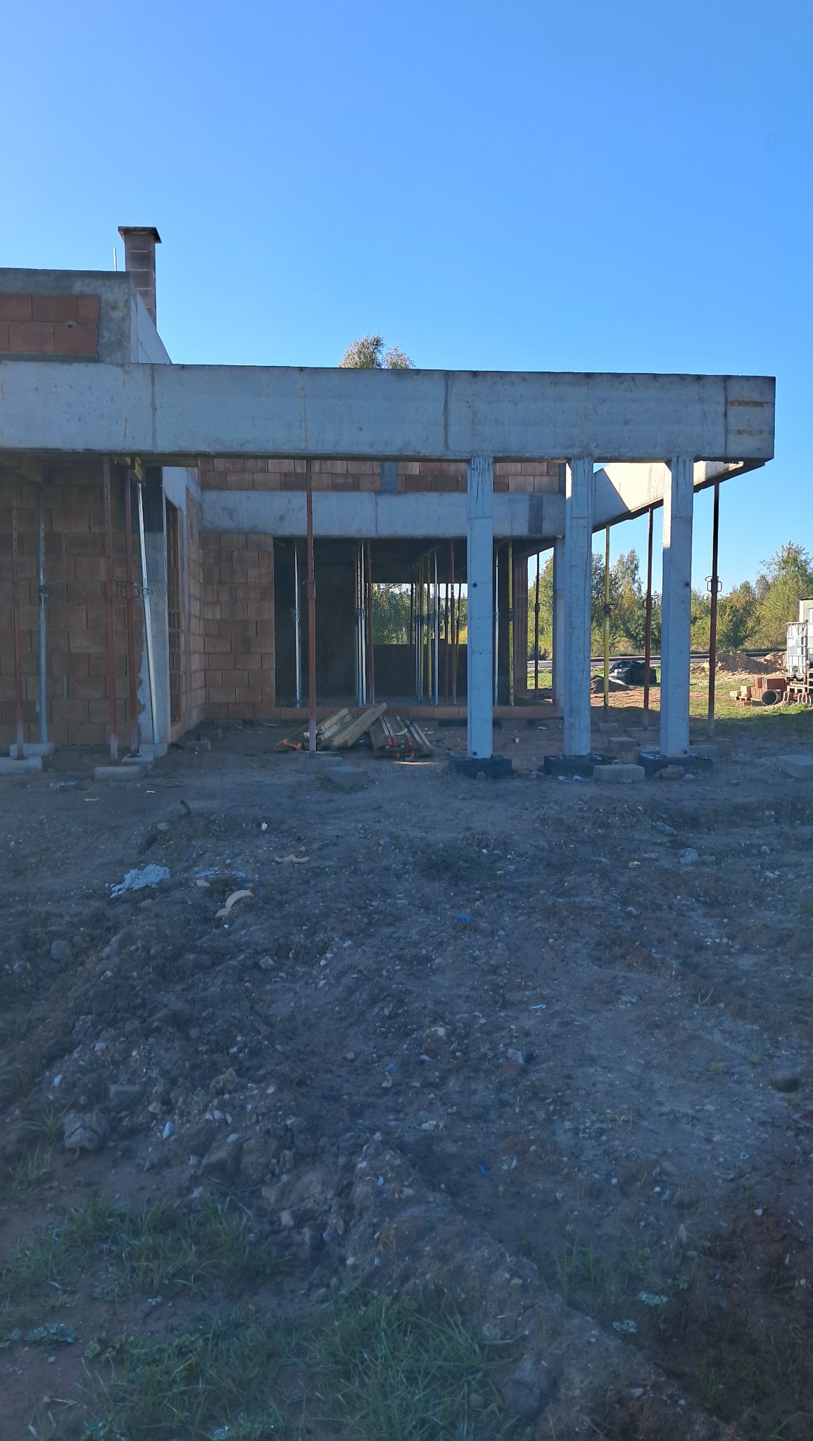 Surowy stan domu murowanego w trakcie budowy: widoczne ściany z cegły, betonowe słupy i strop, teren budowy z ziemią i gruzem, niebieskie niebo w tle.