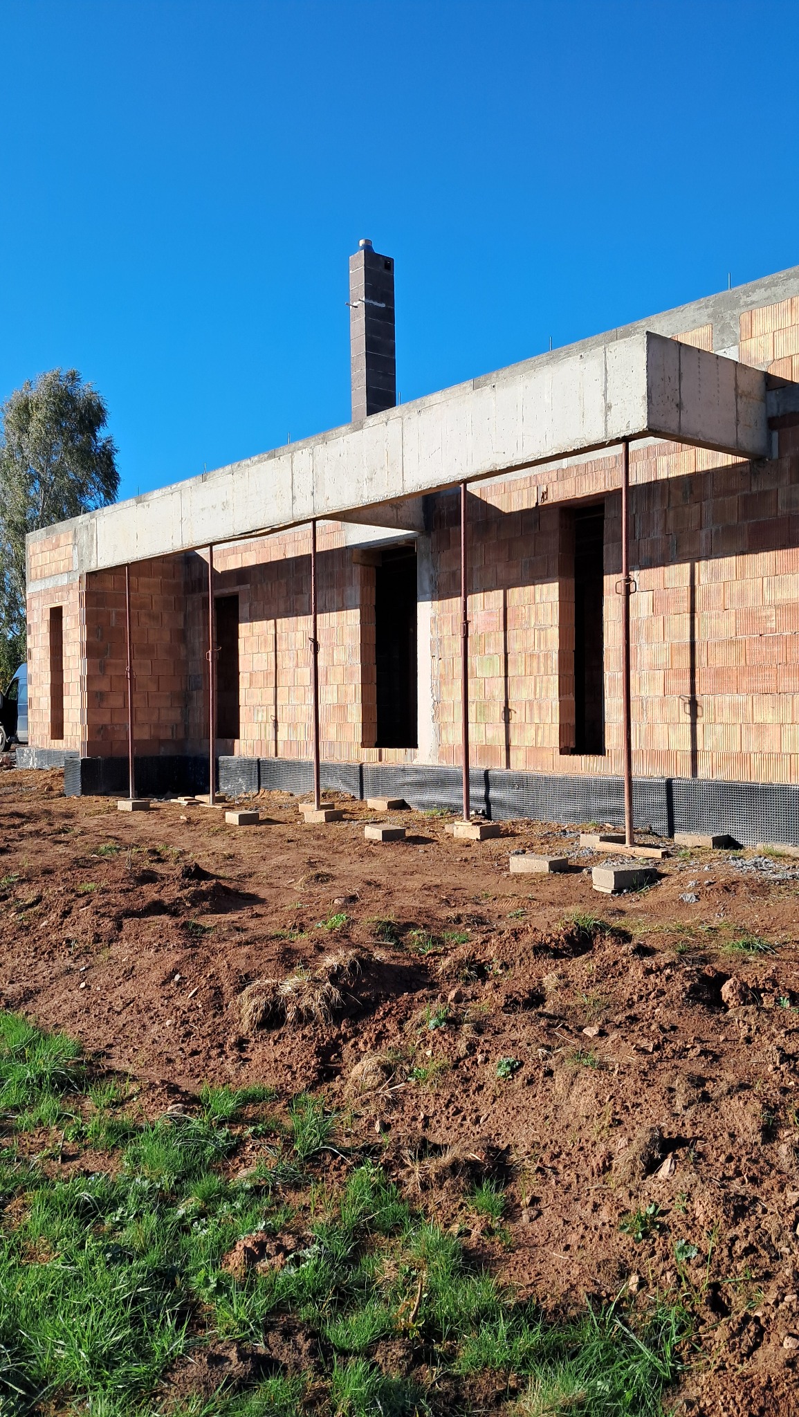 Surowy stan budowy domu z cegły, z widocznym kominem, betonowym wieńcem i metalowymi podporami, na tle błękitnego nieba.